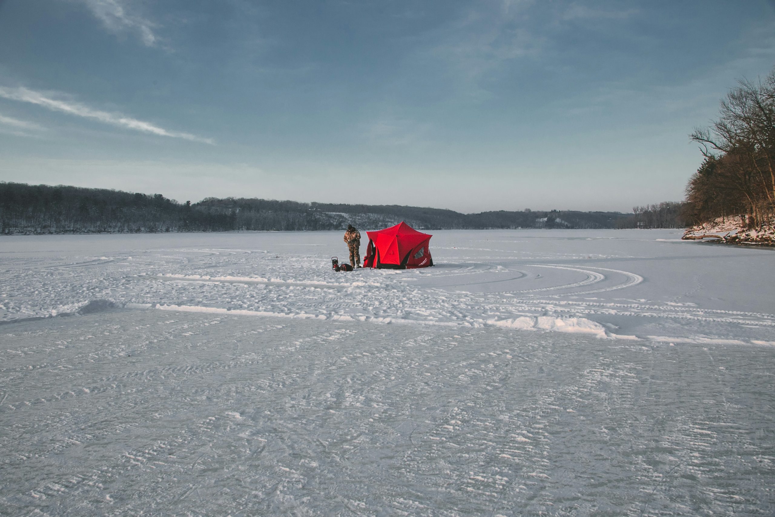 A man standing out on a frozen body of water next to his red ice fishing hut