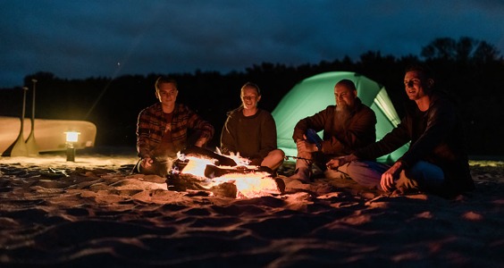 Group of four sitting on a beach in the dark with a bonfire in front of them and a tent set up behind them