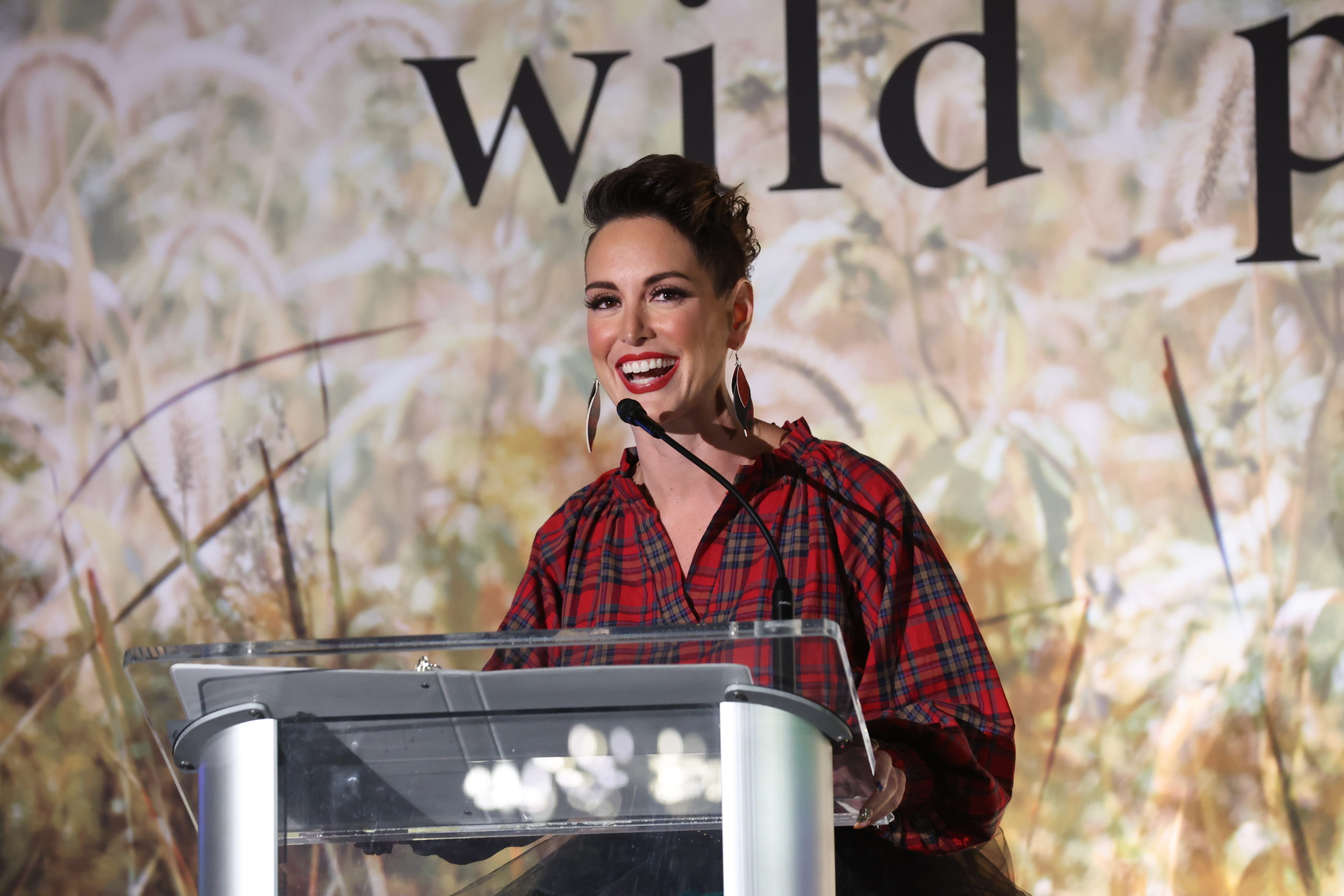 A woman speaking in to the microphone on stage in a red flannel top