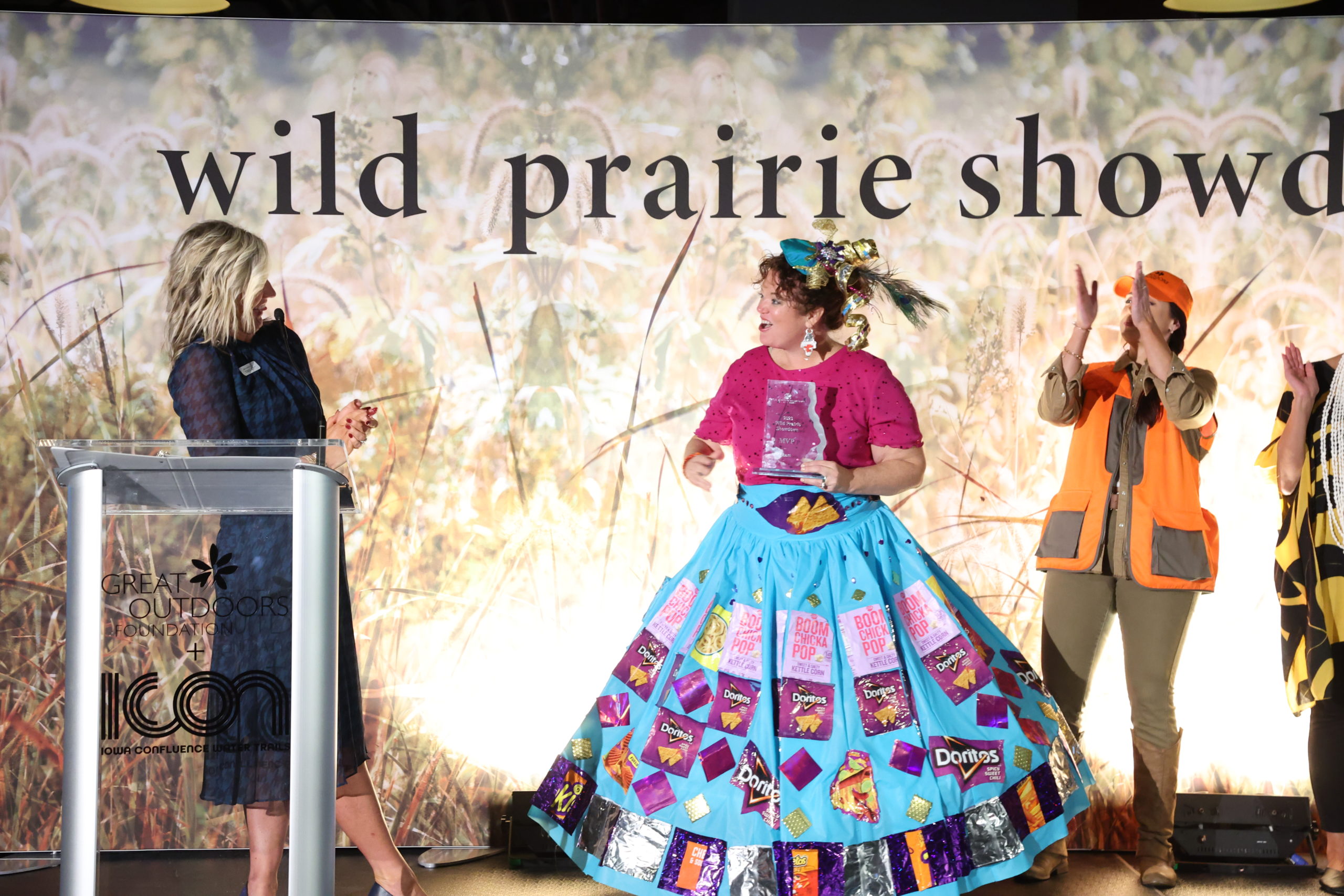 The woman in the skirt with wrapper on it receiving a trophy on stage
