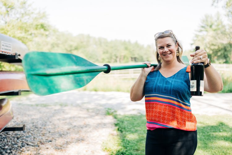 A woman standing next to a stack of canoes and holding a green oar and a dark colored glass bottle