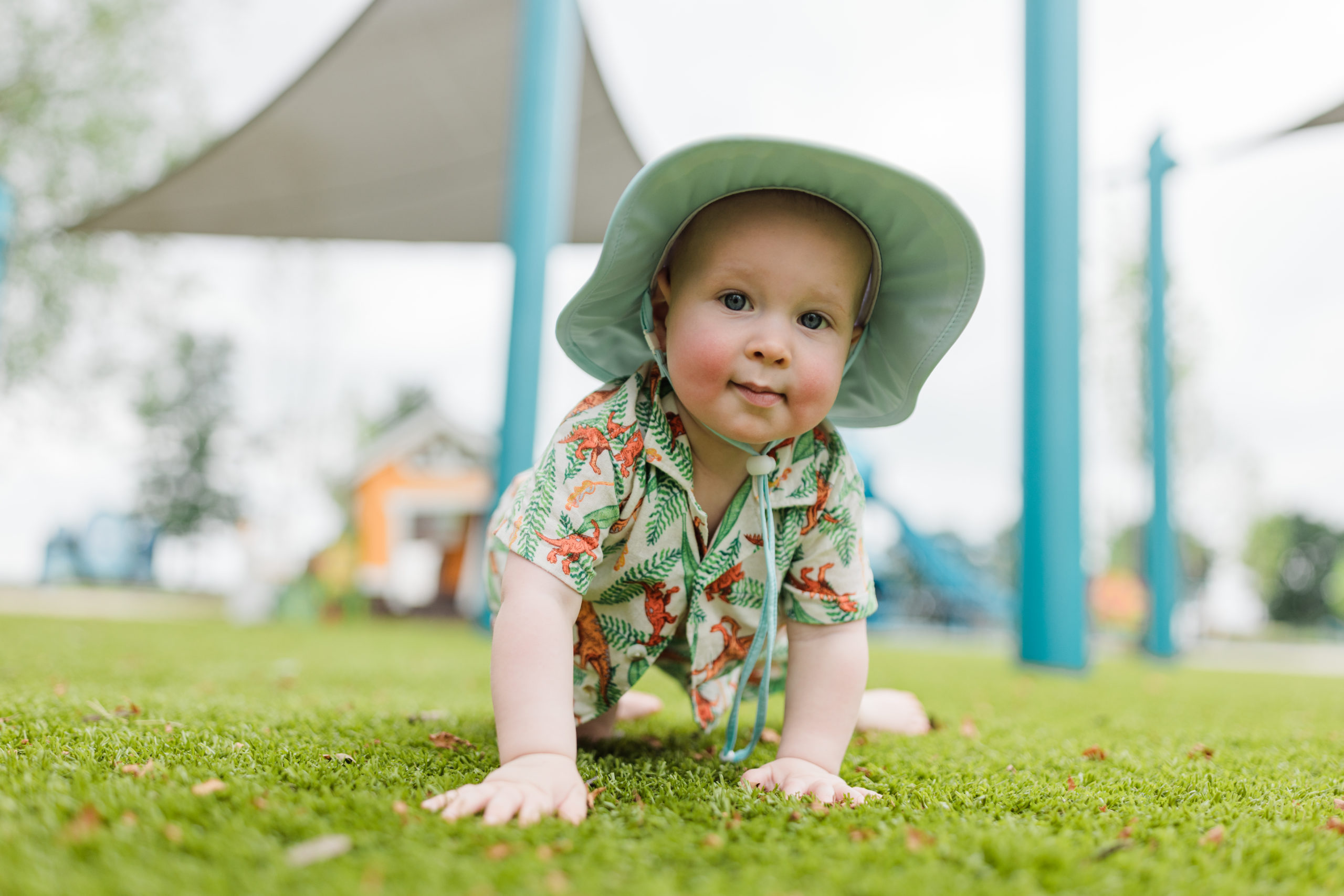 Odin wearing a hawaiian shirt and sunhat while crawling by a playground