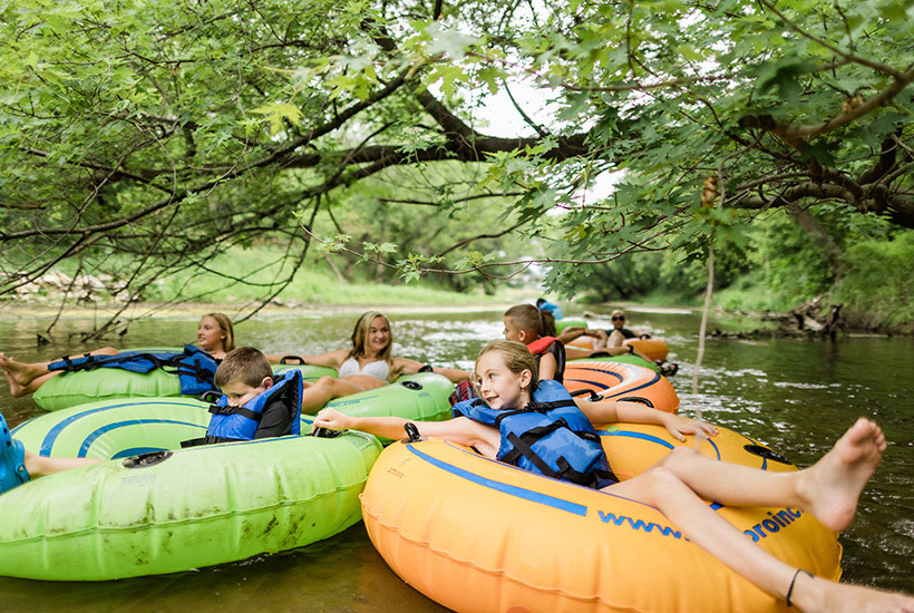A group of kids all holding on to each others tubes as they pass under a branch along the creek