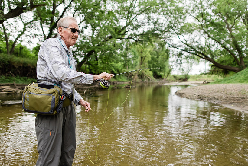 An older gentleman fly fishing from the middle of Beaver Creek. He has a large hip pack and is holding his excess line in his left hand and the fly rod in his right