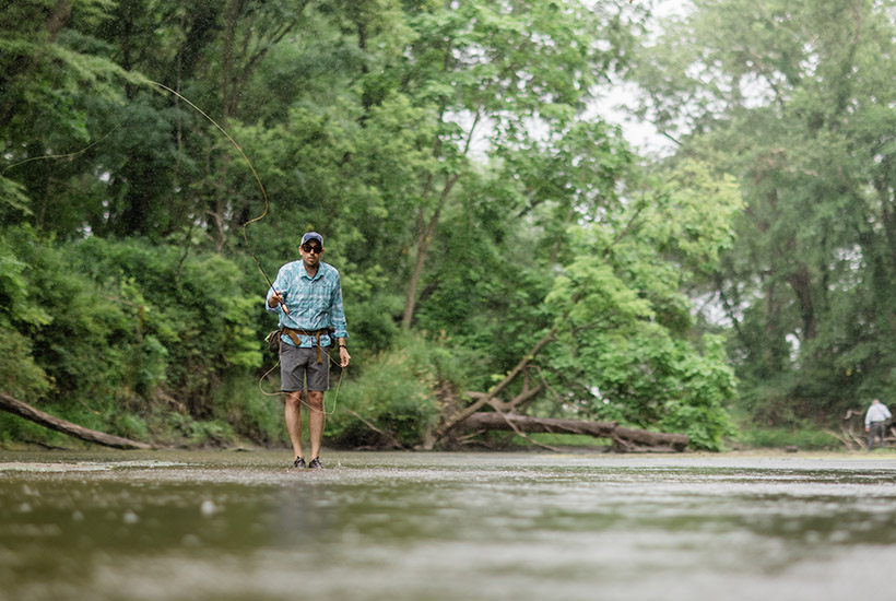 A man mid-cast while fly fishing on Beaver Creek. He's standing in very shallow water and is casting up stream.