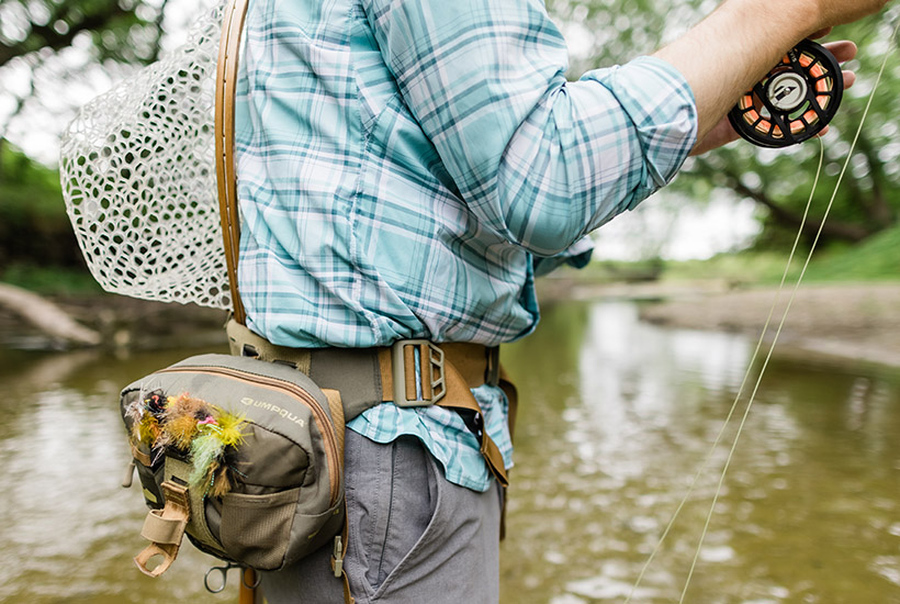 A close up view of a fly fisherman's set up. There are a wide variety of flies attached to his hip pack and he has a net tucked into the back.