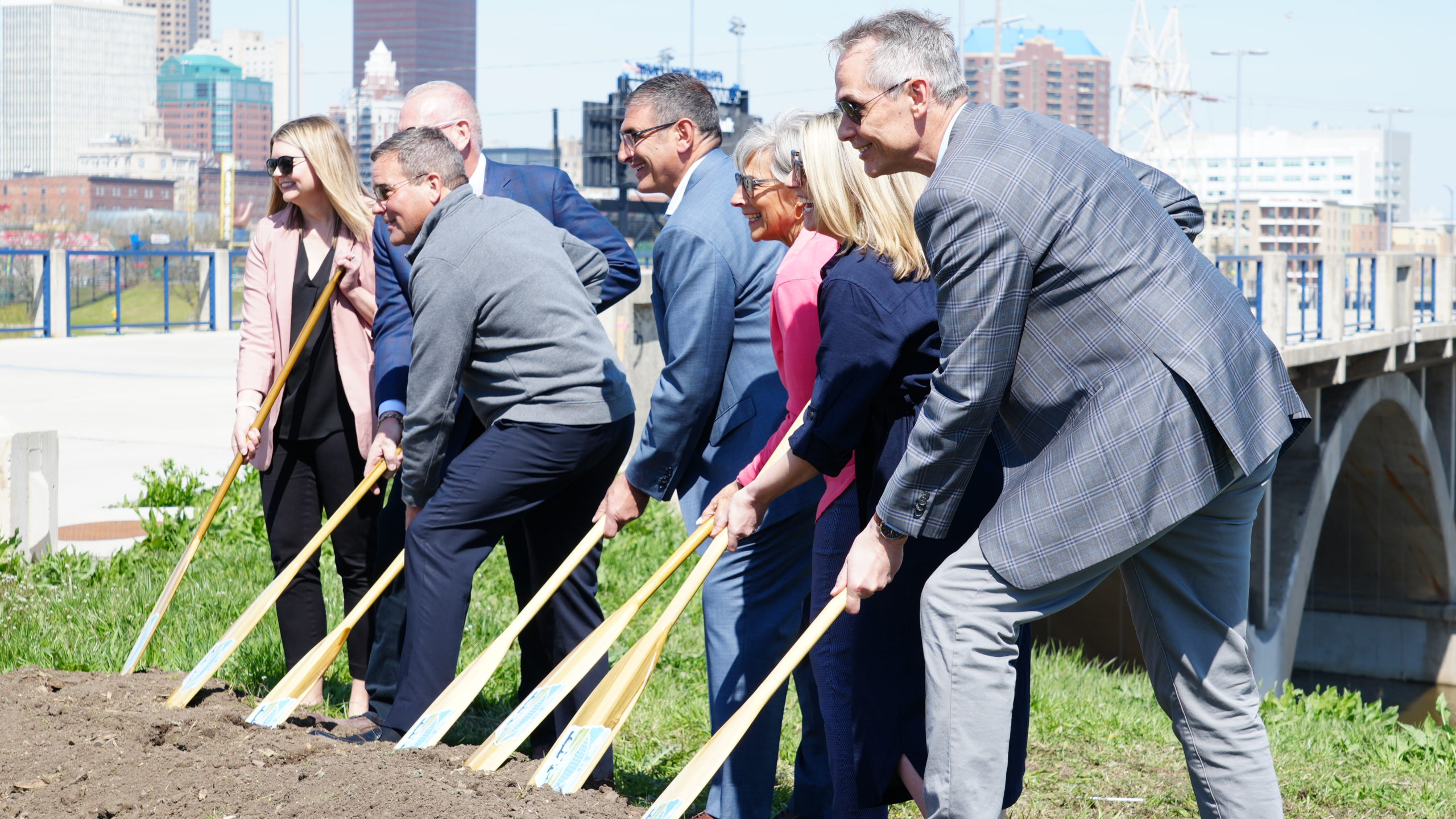 A group of men and women in suits using oars to dig at the groundbreaking ceremony