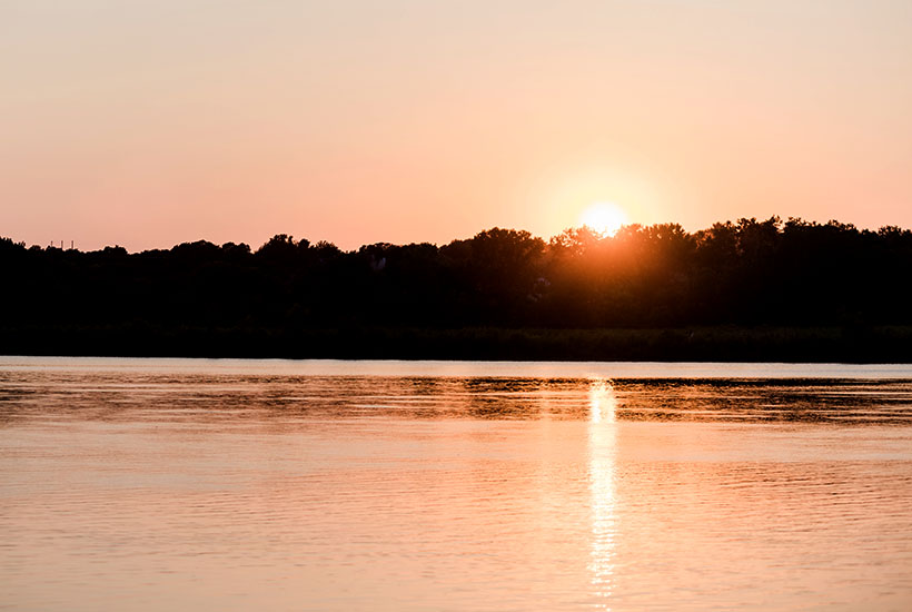 A sunset view of the river. The treeline is completely silhouetted and the top of the sun is poking out above them. The sun is reflecting in the water and the sky and water all have an orange hue