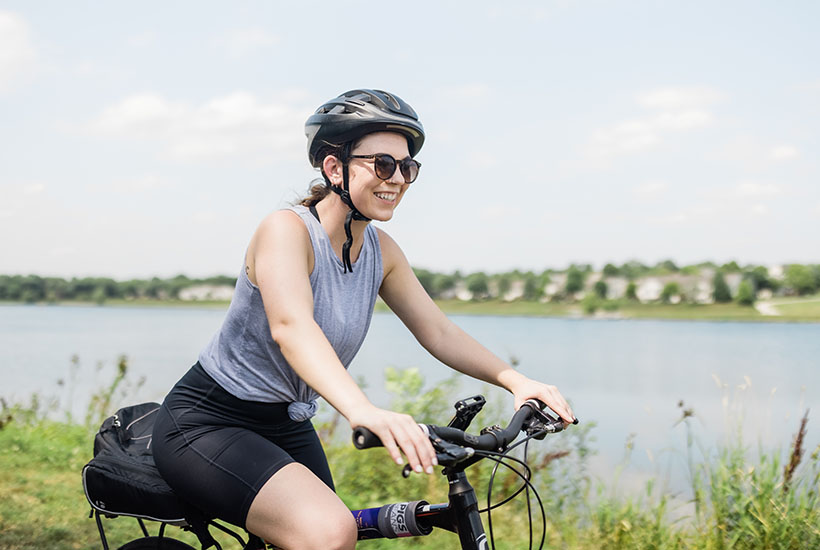 A woman smiling as she rides her bike on a trail next to the river. She's wearing dark shorts, a gray tank top, dark sunglasses and a gray helmet.