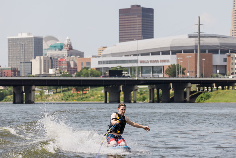A man kneeboarding behind a boat with the Des Moines skyline behind him.
