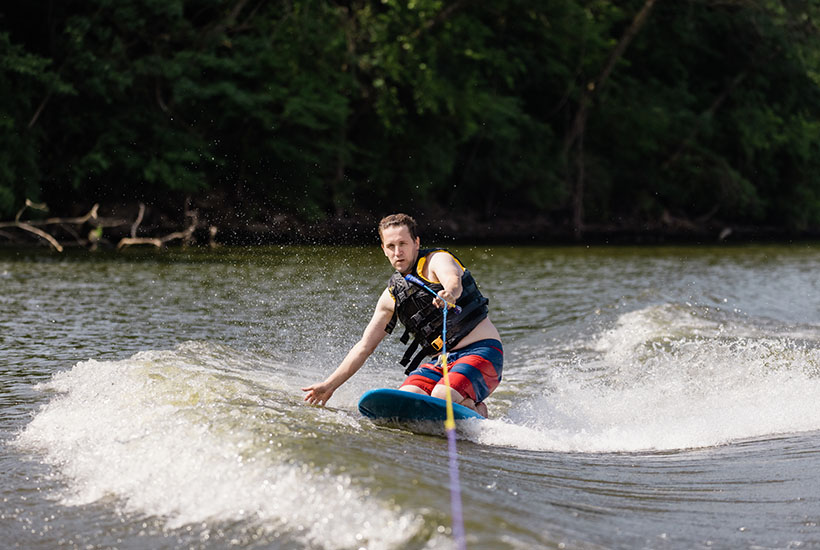 A man kneeboarding along the wake behind a boat. He has one hand on the rope and the other he's dragging in the water next to him while he's turning