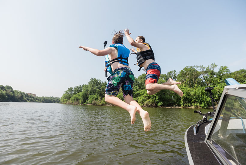 Two people jumping off a boat holding a action camera. Both are wearing swim trunks and lifejackets.