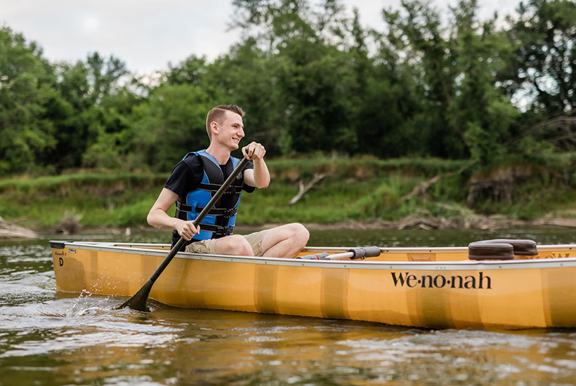A young man smiling as he paddles from the back of the canoe. He's wearing khaki shorts, a dark tshirt and a blue lifejacket.
