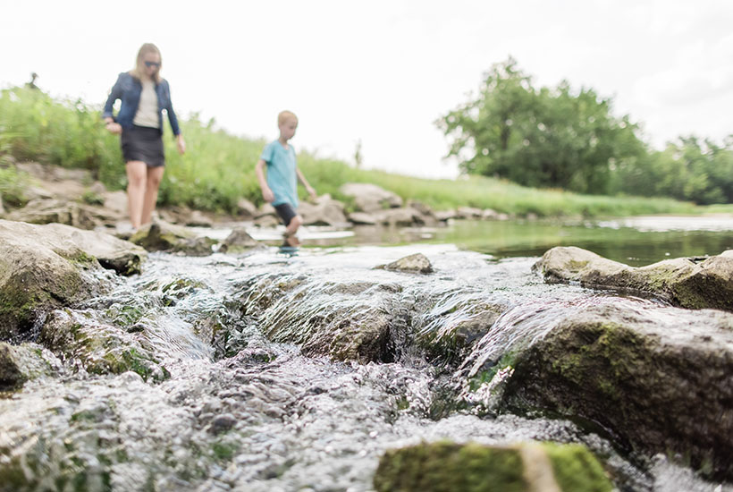 A close up view of water coming over rocks in Fourmile Creek while a mom and her son begin wading in the creek in the distance