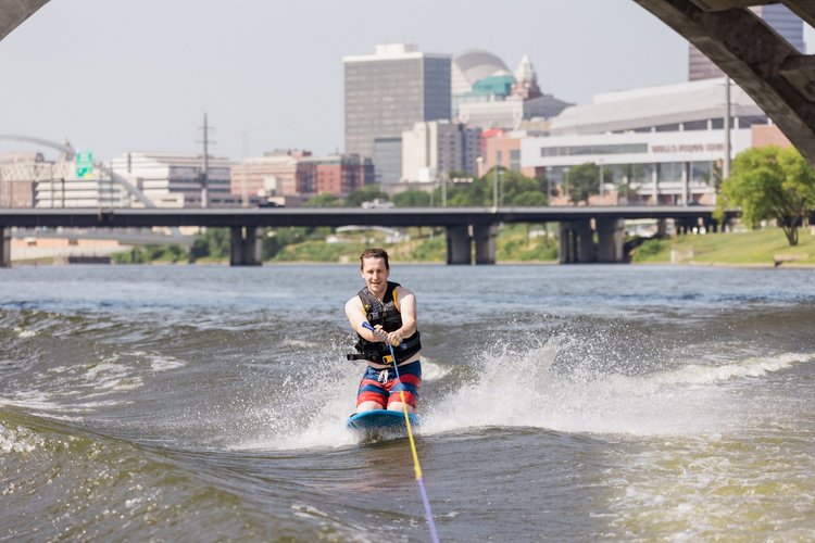 A man kneeboarding with downtown Des Moines behind him. He's smiling and wearing red and blue swim trunks and a black lifejacket