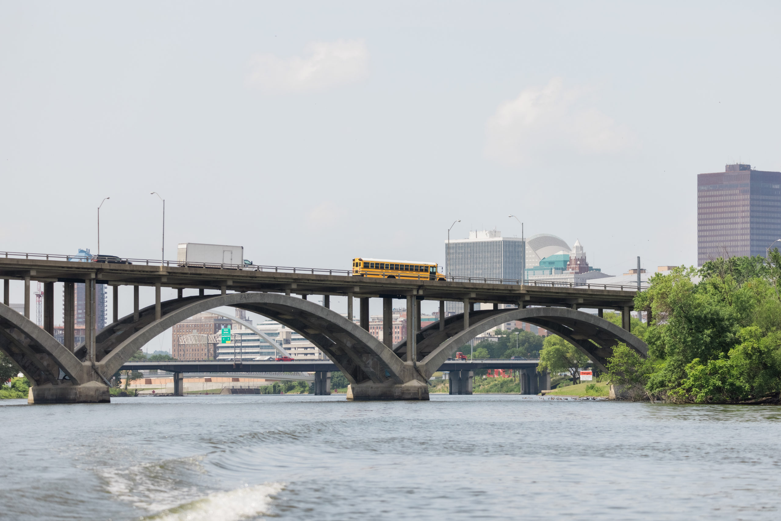 A view from the river looking up at a downtown bridge with a school bus and other vehicles crossing over it