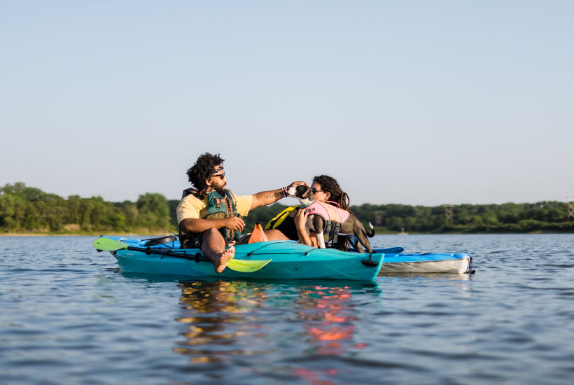 Two people taking a break while they kayak. The man has his feet dangling out the sides of the kayak and is reaching over and petting a brown and white dog that's standing on the back of the other kayak.