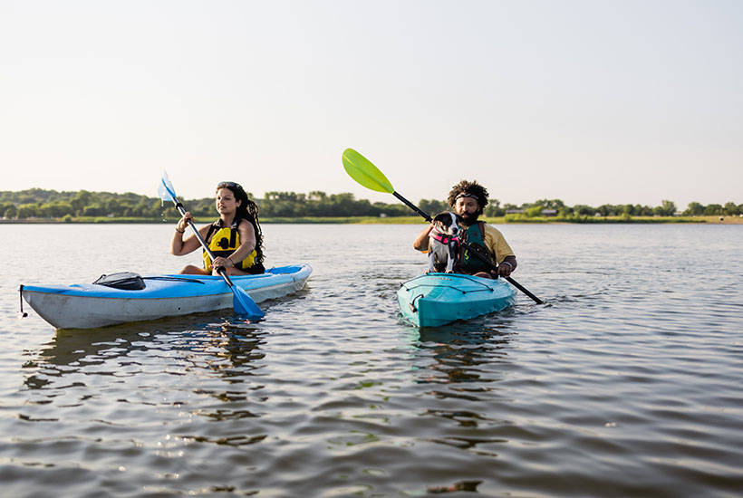 A man and a woman kayaking - they're both in blue kayaks and wearing lifejackets. The man has a dog sitting in his that's also wearing a lifejacket.
