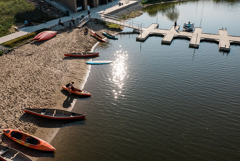 A beach with canoes and kayaks along it. There's a woman getting her lifejacket out of a kayak and another person on the dock walking towards a boat that's tied up