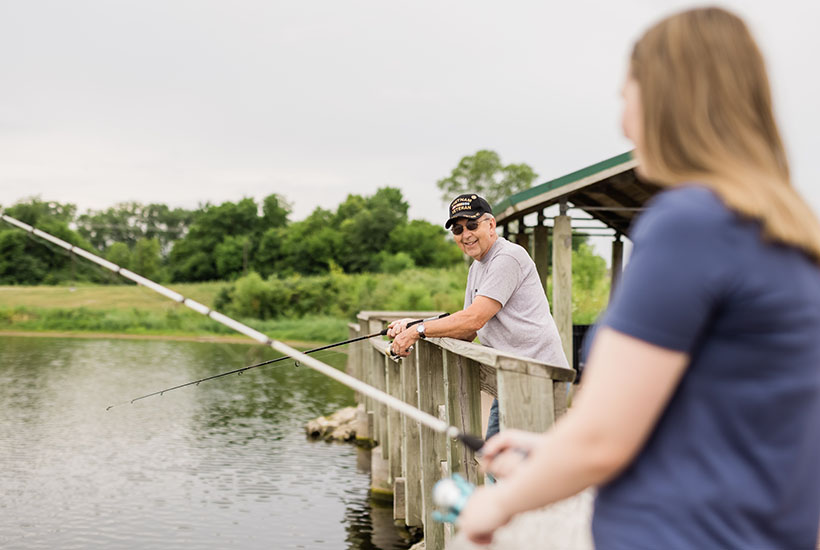 An older gentleman with a Veteran hat fishing from the pier and smiling towards his daughter.