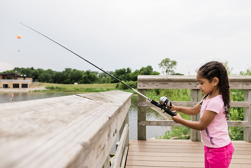 A little girl in a pink outfit holding a fishing pole above the water after reeling the line in.