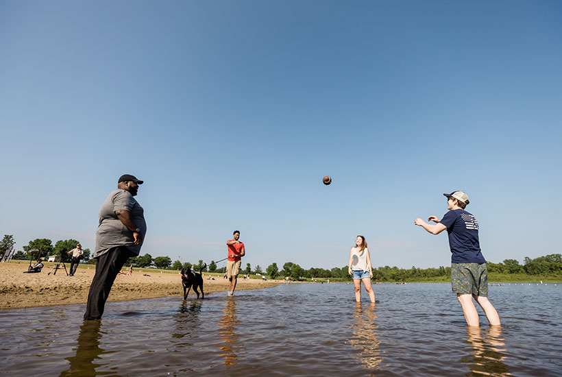 A group of four people standing in shallow water next to the beach and playing catch with a football.