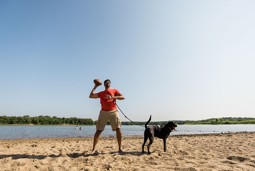 A man getting ready to throw a football while holding his dog's leash on a beach