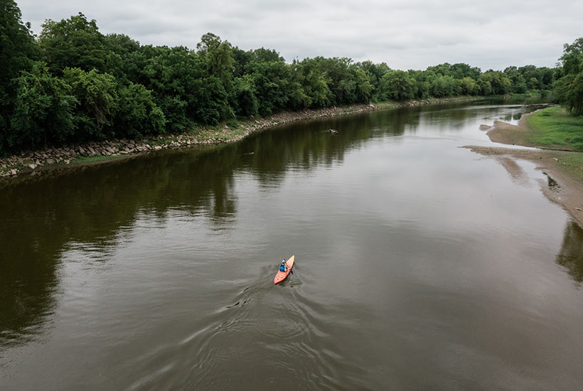 An above view of a kayaker going down the river. The river is tree lined and you can see a sand bank on the right side
