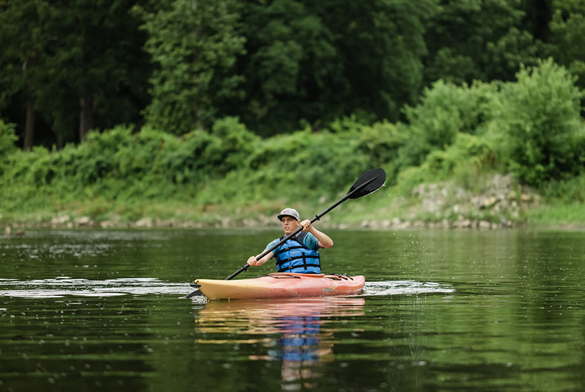 A man paddling in a kayak with green trees and shrubbery behind him on the coastline.