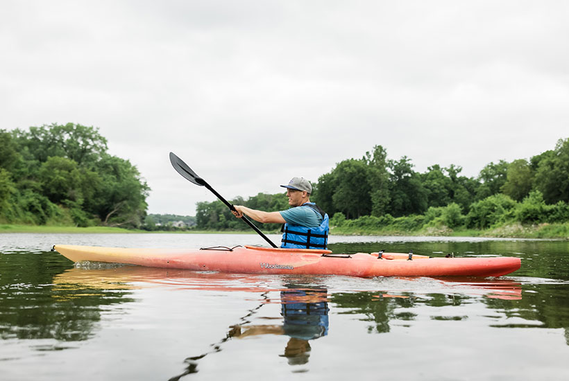 Side view of a kayaker in the Raccoon River. The kayak is orange and yellow and the man in the kayak is wearing a blue life jacket and a baseball cap.