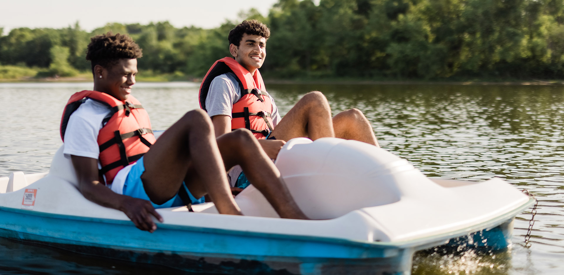 Two young men in a blue and white rental pedal boat wearing orange life jackets