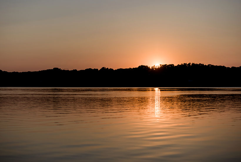 A sunset over a body of water. The top of the sun poking over the treeline is reflected in the water and the water and sky are both an orange hue