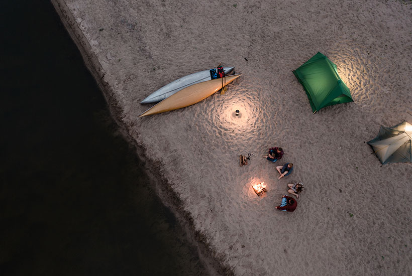 An above view of a camp set up on the sand next to the river. There are two canoes, two tents and a group of four people sitting near a fire.