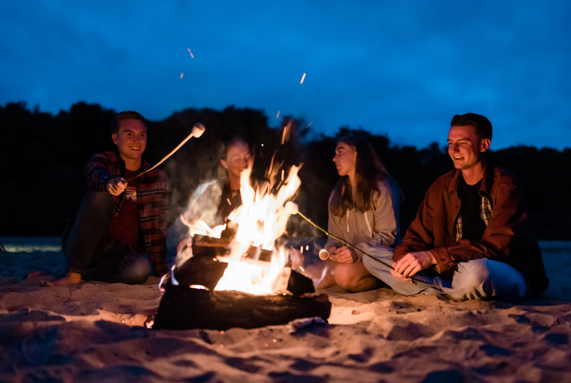 A group of four sitting around a campfire on a sandbar and roasting marshmallows.