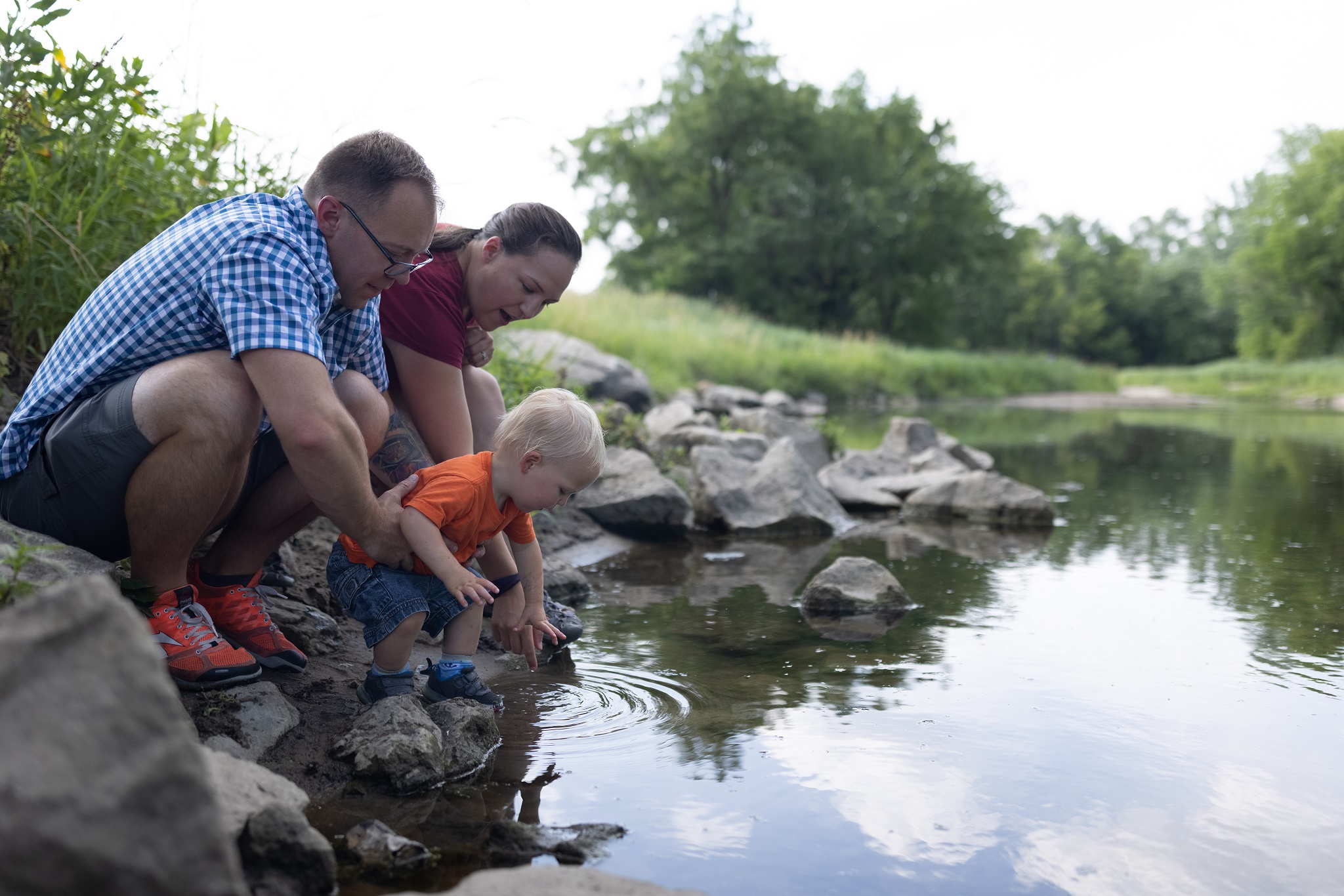 2X5A1080 Young family helping their toddler touch the water in a stream